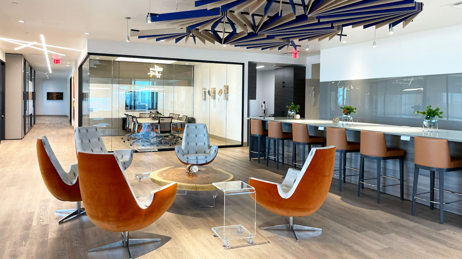A modern office lounge area features four orange swivel chairs around a round wooden coffee table, a clear acrylic side table, a long counter with brown bar stools, and a glass-walled conference room in the background.